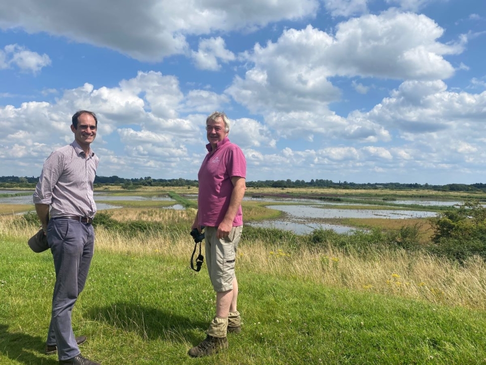 Two men standing on a grassy river back overlooking recreated wetlands under a blue sky with patchy clouds.